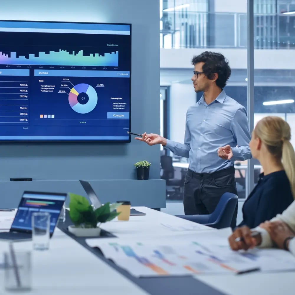 A presenter pointing at a wall screen of charts in a glass meeting room, colleagues at the table