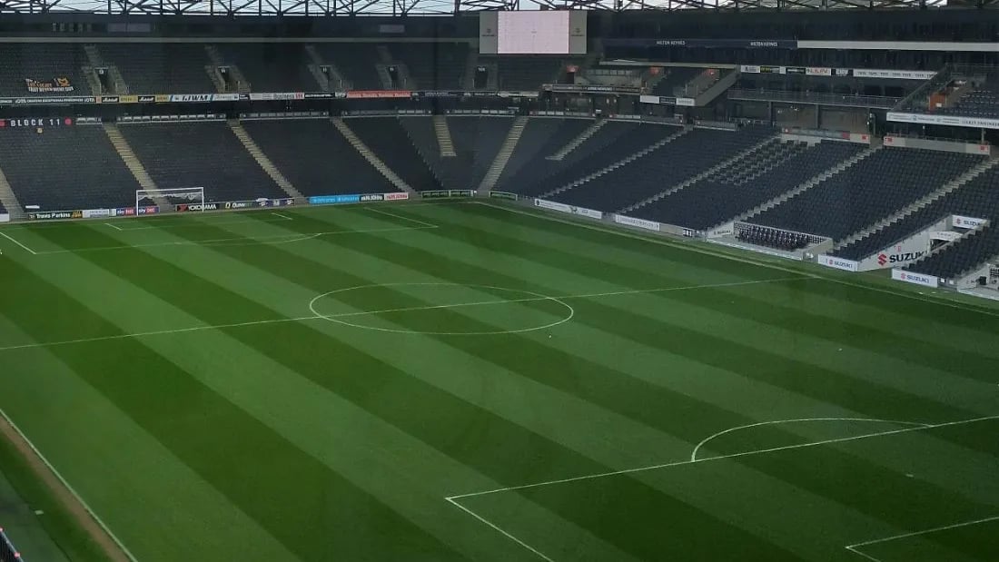An empty football stadium from the upper tier, stripes mown into the pitch and the far stand in shade