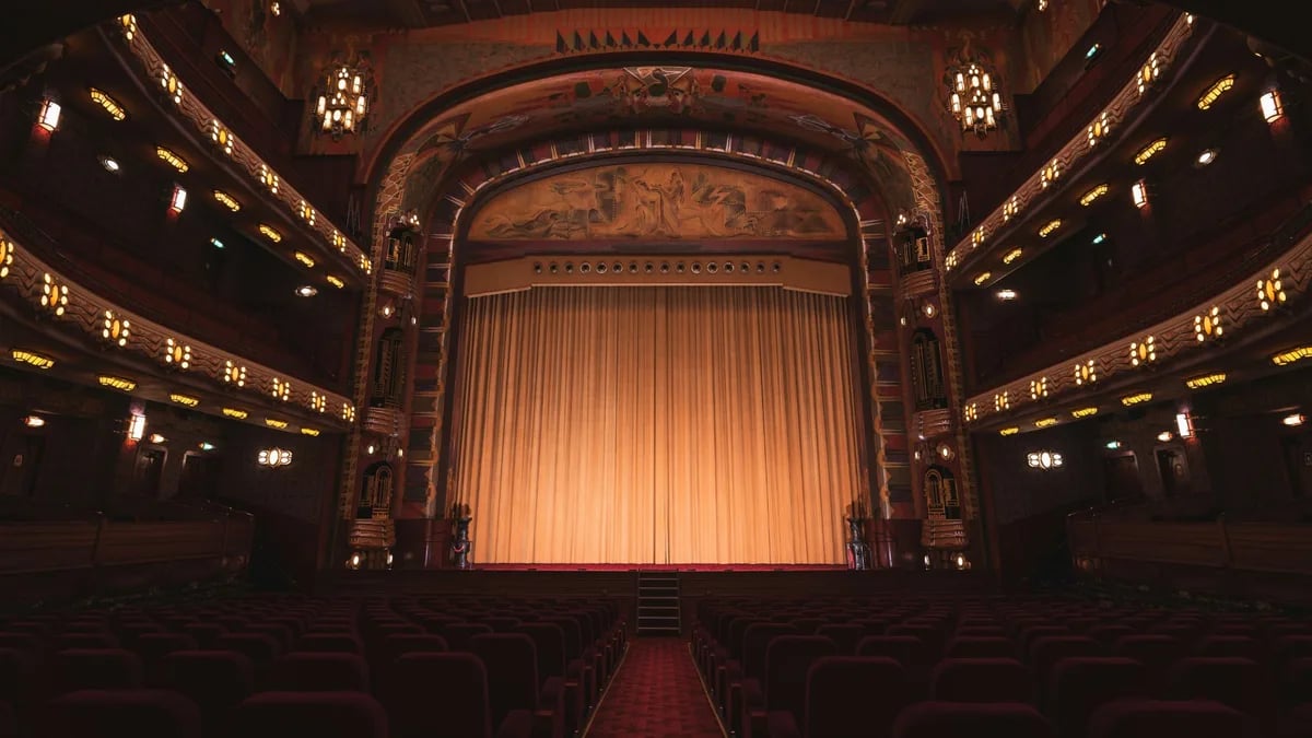 An ornate theatre auditorium from the stalls, red velvet seats and a lit curtain across the stage
