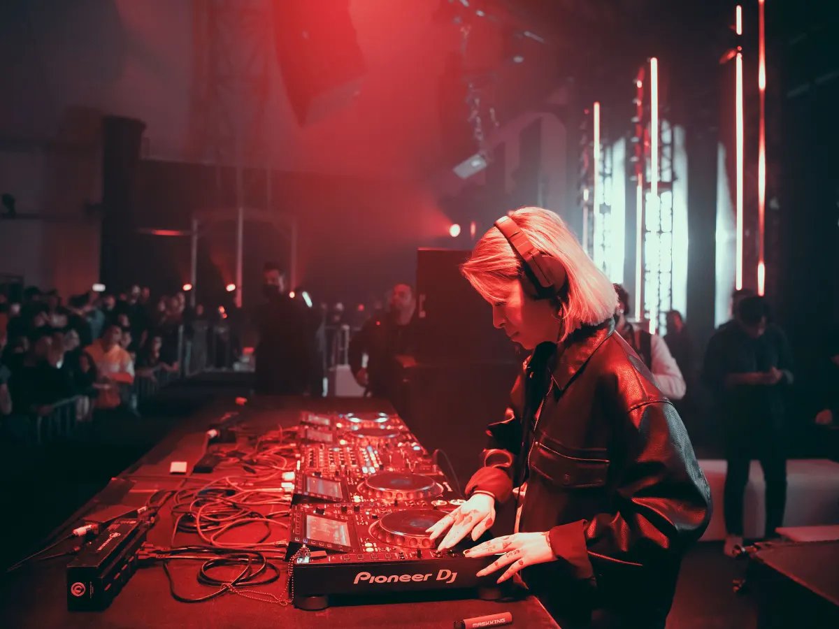 A DJ in headphones at the decks under red light, the crowd behind the barrier