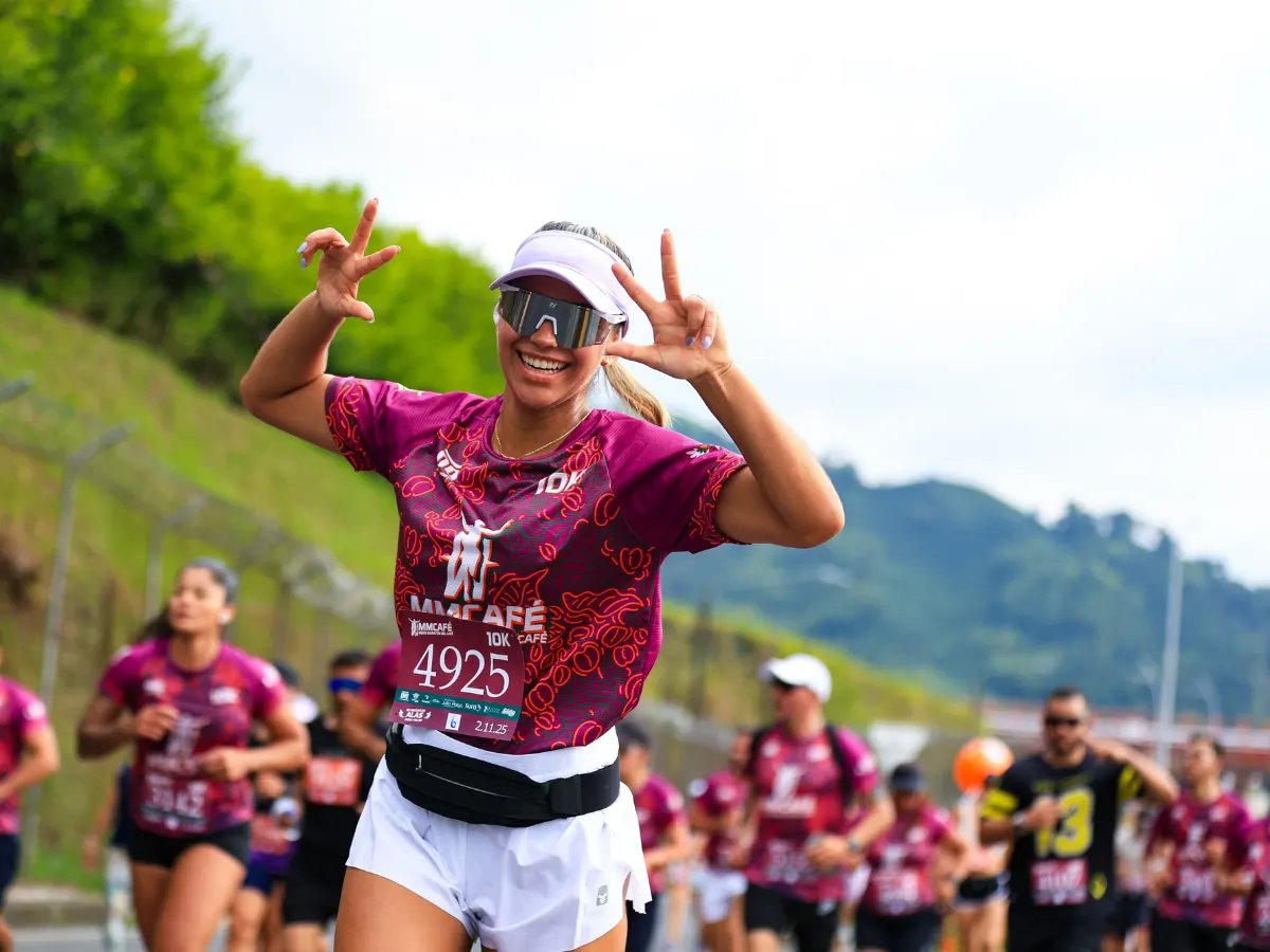A runner in a race bib throwing peace signs mid-race, a pack of runners behind her on a hillside road